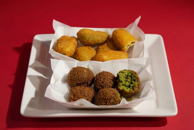 Two white paper trays on a white plate, one with golden fried jalapeño poppers, the other with falafel.