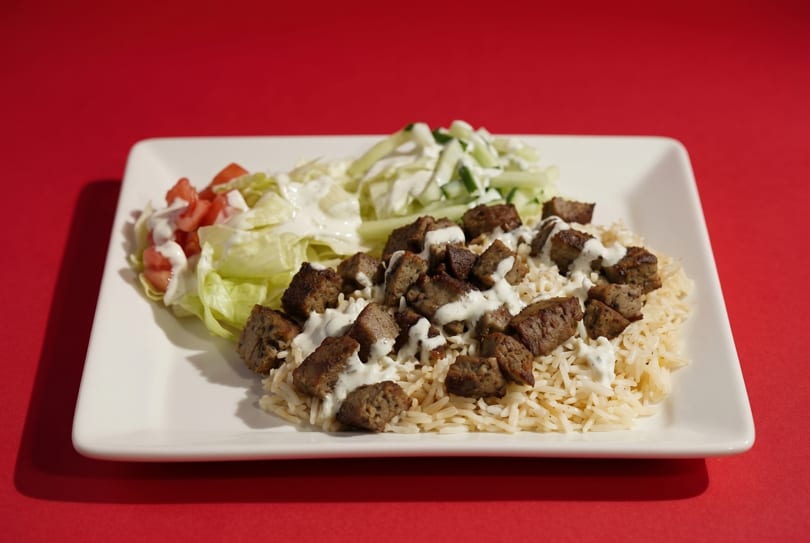 A white rectangular plate holds rice, meat, and two salads, drizzled with white sauce on a red background.